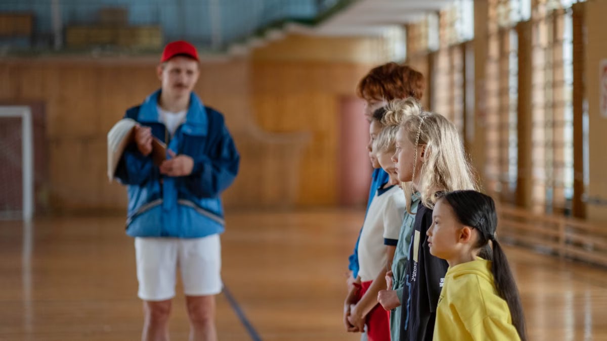Diverse group of children standing indoors in a gym with a coach in focus.