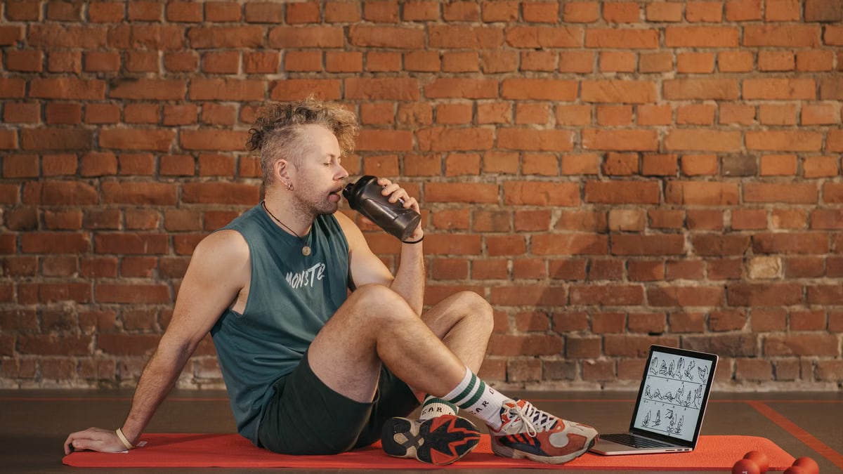 Athletic man sitting on exercise mat with water bottle, taking a break from workout, in a rustic indoor setting.